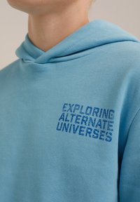 Vaaleansininen huppari, jossa on pehmeä pinta, ja edessä vasemmalla puolella on tummansinisellä teksti "TUTKIMASSA ALTERNATIIVISIA UNIVERSUMEJA".