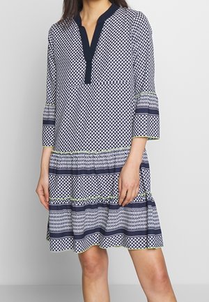 Kobieta w sukience do kolan z geometrycznymi wzorami w kolorach granatowym i białym, z rękawami do połowy przedramienia i granatowym kołnierzem w kształcie litery V.