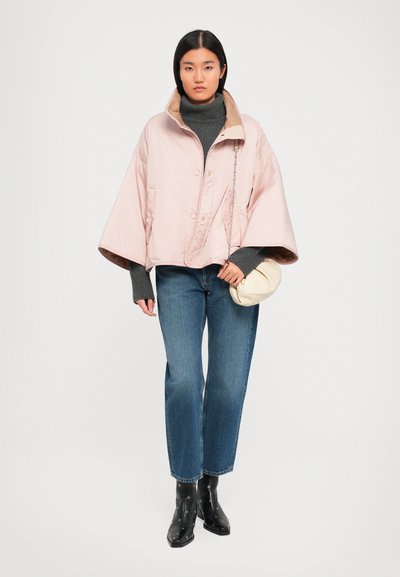 Žena v svetloružovej oversized bunde, šedom roláku, modrých džínsoch, čiernych členkových čižmách, drží malú krémovú kabelku.