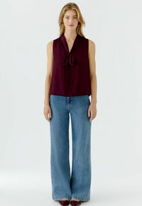 Bordeauxfarbene ärmellose Bluse mit einem gebundenen Vorderteil, kombiniert mit hochgeschnittenen, weit geschnittenen blauen Jeans. Glatter Stoff und lockere Passform.