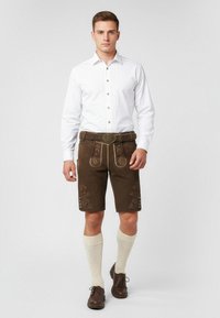 Lederhosen marroni con motivi floreali ricamati, abbinate a una camicia bianca con bottoni e calze crema al ginocchio, accentuate da una fibbia rotonda della cintura.