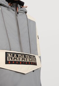 Grå och krämfärgad jacka med dragkedja fram, huva och orange dragsnören. Har en framträdande svart och orange logotypapplikation.