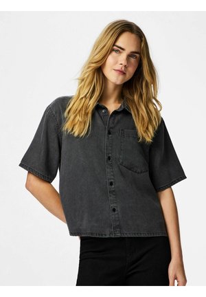 Jeune femme aux cheveux longs et blonds, portant une chemise ample à manches courtes gris foncé boutonnée et un pantalon noir, posant devant un fond uni.