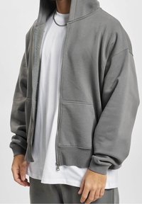 Sweat à capuche gris zippé avec une capuche, des poches frontales et des poignets côtelés. Porté sur un t-shirt blanc, avec une texture lisse et une coupe décontractée.