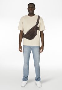 Borsa a tracolla in pelle marrone indossata a tracolla, dotata di zip, tracolla regolabile e un dettaglio in metallo circolare. Modello con camicia chiara e jeans.