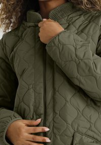 Chaqueta acolchada verde oliva con cuello acanalado, cierre de cremallera y patrón de costura curvada. Tejido texturizado con bolsillos laterales.