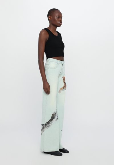 Zwarte geribbelde crop top, licht denim wijde jeans met katten- en kittenprints, gladde textuur, contrasterende kleuren, platte schoenen, model geposeerd zijwaarts.
