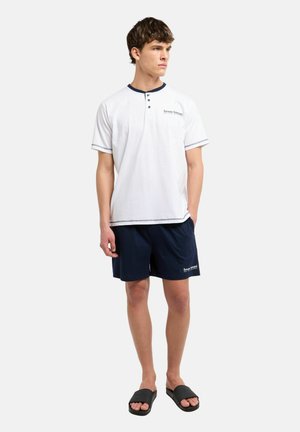 Jonge man die een wit henley shirt met korte mouwen, marineblauwe korte broek en zwarte slides draagt, staand met één hand in de zak.