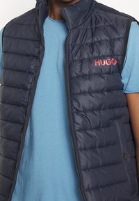 Navyblaues, gepolstertes Weste mit hohem Kragen, Reißverschluss vorne und horizontalem Steppmuster. Mit rotem "HUGO"-Logo auf der Brust.