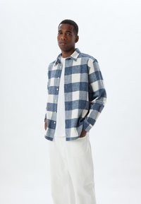 Camicia di flanella a quadri blu e bianchi con design a bottoni, abbinata a pantaloni bianchi con texture e una semplice t-shirt bianca sotto.