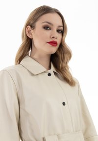 Cappotto in pelle beige chiaro con colletto classico, dotato di bottoni neri e vita segnataspettatore. Tessuto liscio con una vestibilità su misura.