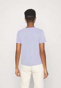 Fialové bavlněné tričko s krátkými rukávy, kulatým výstřihem a hladkou texturou. Nosené s světlými pruhovanými kalhotami. Pohled zezadu.