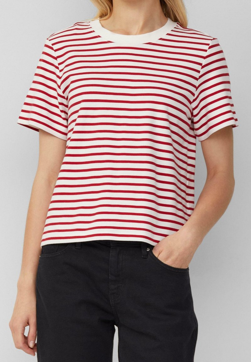 Femme portant une chemise blanche à manches courtes avec des rayures rouges horizontales et un pantalon noir, une main dans la poche, sur fond gris clair.