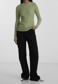 Femme portant un pull à manches longues côtelé vert clair, un pantalon large noir et des chaussures métalliques pointues, debout avec une main dans la poche.