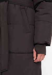 Καφέ παλτό με επένδυση, με ζώνη στη μέση, πλαϊνές τσέπες και μανσέτες με τρύπα για τον αντίχε�ιρα. Λείο, ματ ύφασμα με ραμμένες ζώνες για μόνωση.