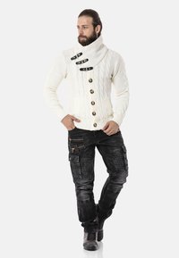 Fülliger Cream-Kabelstrickpullover mit hohem Kragen, versehen mit vier schwarzen Toggle-Knöpfen. Kombiniert mit dunklen Cargo-Hosen mit Reißverschluss-Akzenten.