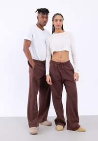 Twee modellen dragen casual outfits; de een in een wit t-shirt met korte mouwen, de ander in een cropped ribbed top, gecombineerd met bruine joggers en beige schoenen.