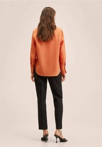 Camisa de mangas largas naranja con puños abotonados, combinada con pantalones negros ajustados y tacones negros de tiras, vista desde la parte trasera.