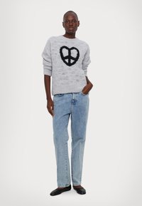 Ανοιχτό γκρι πλεκτό πουλόβερ με σχέδιο μαύρης ειρηνικής σημαίας, συνδυασμένο με ανοιχτό μπλε τζιν παντελόνι και μαύρα παπούτσια. Υφή λεπτομέρειας στο πουλόβερ.