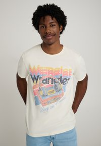 Cream t-shirt with retro graphic print featuring a cassette player and "Wrangler" text in bold colors. Short sleeves and a round neckline.