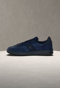 Σκούρο μπλε sneaker χαμηλής κοπής με κορδόνια, μείγμα σουέντ και υφάσματος, και μαύρη λαστιχένια σόλα σε ανοιχτή επιφάνεια με γκρι φόντο.