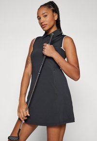 Svart ärmlös golfklänning med krage, som har vita detaljer, en figursydd silhuett och en slät tygtextur. Håller en golfklubba.