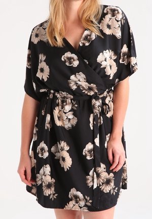 Sort omslagskjole med beige og hvidt blomsterprint, korte ærmer og et bånd i taljen, båret af en stående person.