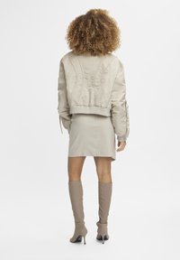 Vaalean beige-tyyppinen bomber-takki, jossa on ribbireunat, yhdistettynä istuvaan beigeen mini-hameeseen ja polveen asti ulottuviin beigeihin saappaisiin, luoden monochromaatista ilmettä.