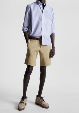Homme portant une chemise à manches longues rayée bleue, un short beige avec une ceinture marron et des chaussures à lacets beiges, debout devant un fond uni.