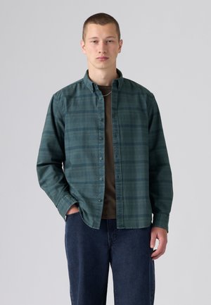 Jeune homme debout, une main dans la poche, portant une chemise à carreaux vert foncé boutonnée sur un t-shirt marron et un jean bleu, sur un fond gris.