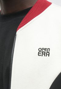 Chaqueta negra y blanca con cremallera, que presenta un cuello rojo acanalado y un logo bordado "OPEN ERA" en el panel lateral blanco. Textura suave.