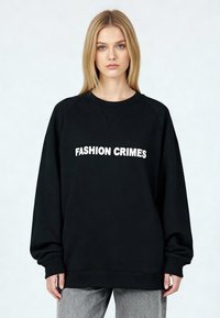 Sweat-shirt noir avec le texte "FASHION CRIMES" en blanc, doté d'un col rond, de manches raglan longues et de poignets et ourlet côtelés.