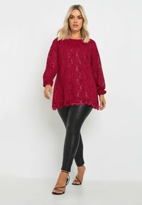 Top in pizzo rosso con maniche lunghe, orlo ondulato, indossato sopra leggings neri in finta pelle e tacchi neri con cinturini.