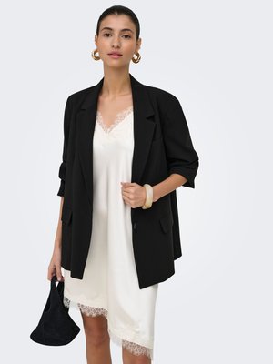 Femme portant une robe nuisette blanche bordée de dentelle sous un blazer noir, tenant un petit sac à main noir, accessoirisée avec des boucles d'oreilles créoles dorées et un bracelet.