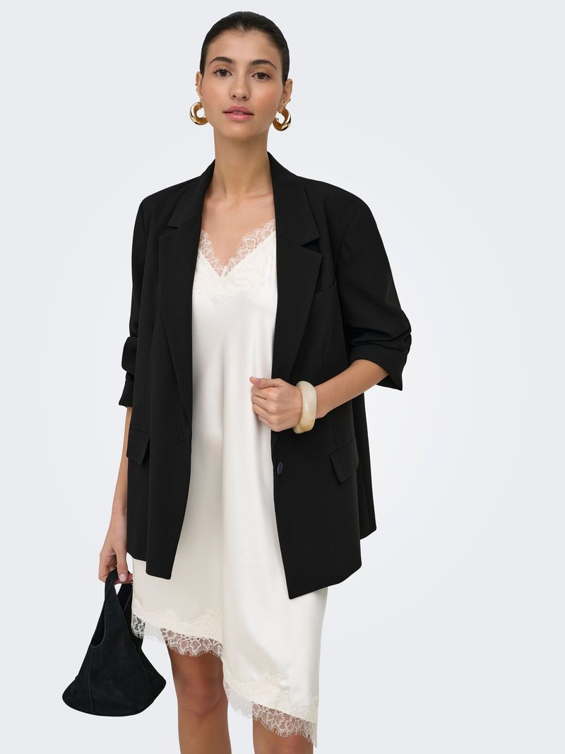 Femme portant une robe nuisette blanche bordée de dentelle sous un blazer noir, tenant un petit sac à main noir, accessoirisée avec des boucles d'oreilles créoles dorées et un bracelet.