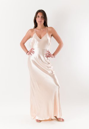 Kobieta stoi z rękami na biodrach, ubrana w jedwabną, jasnoróżową sukienkę do ziemi i sandały na otwartym obcasie na jednolitym białym tle.