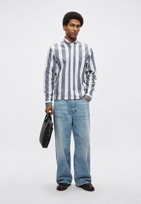 Ανδρικό πουκάμισο με κουμπιά με κάθετες γκρι και άσπρες ρίγες, συνδυασμένο με ανοιχτό μπλε τζιν παντελόνι με φαρδύ πόδι. Κρατάει μια μαύρη τσάντα, στέκεται σε απλό φόντο.