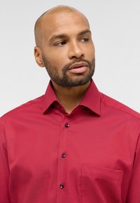 Camicia rossa con bottoni, realizzata in un tessuto uniforme, dotata di una tasca sul lato sinistro del petto e bottoni coordinati. Design con colletto classico.