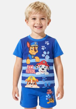 Garçon blond souriant portant un t-shirt bleu Paw Patrol et un short avec des chiens dessinés et des empreintes de pattes.