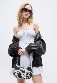 Νεαρή γυναίκα με μαύρο δερμάτινο μπουφάν που πέφτει από τους ώμους, λευκό φόρεμα με ρυτίδες, σκούρα γυαλιά ηλίου, κρατώντας τσάντα με ασπρόμαυρο μοτίβο.