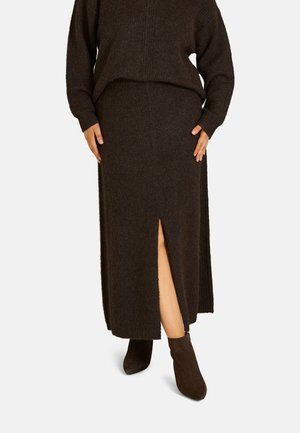 Femme portant une longue jupe en maille marron foncé avec une fente devant, assortie à un pull en maille et des bottines marron.
