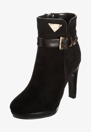 Stivaletto alla caviglia in suede nero con tacco alto, cinturino nero lucido con fibbia dorata e targhetta triangolare dorata con il logo sulla parte esterna superiore.