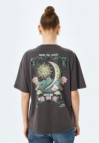 Grijze oversized t-shirt met een achterafbeelding van een zon, halve maan, bloemen en paddenstoelen. Korte mouwen en zachte katoenen stof.