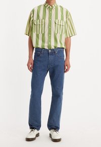 Grön och krämrandig kortärmad skjorta med två bröstfickor, kombinerad med blå raka jeans och vita snörskor.