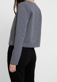 Chaqueta de tela gris con relieve, de diseño cropped y bolsillos laterales con cremallera. La parte trasera es lisa y tiene mangas largas para mayor cobertura.
