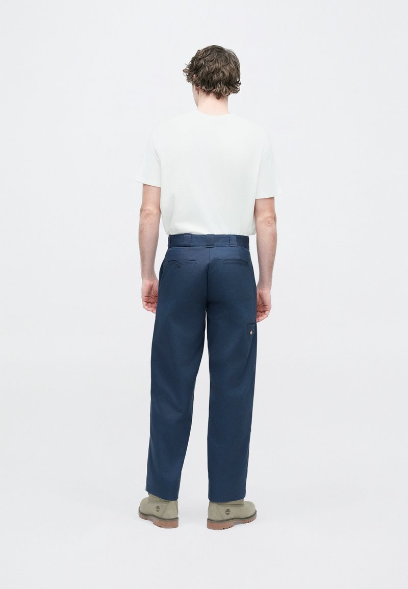 Pantalones azul marino de corte recto, con un detalle de bolsillo trasero y costuras sutiles, combinados con una camiseta blanca y calzado gris.