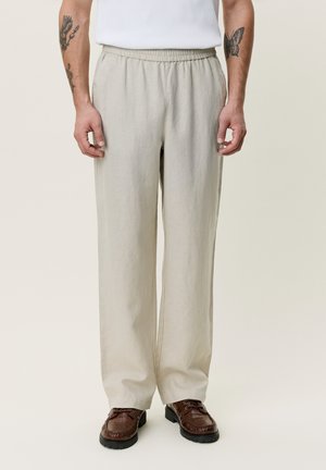 Man draagt beige broek met wijde pijpen en elastische taille en bruine leren schoenen, staand tegen een neutrale achtergrond.