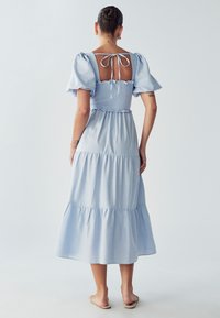 Svetlo modra, večslojna maksi obleka z napihnjenimi rokavi, smockanim korzetom in odprtim hrbtom. Vključuje detajl s trakom in mehko teksturo tkanine.