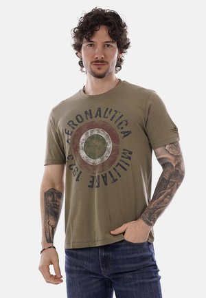 Uomo con capelli ricci che indossa una t-shirt verde oliva Aeronautica Militare e jeans, mostrando tatuaggi su entrambi gli avambracci, mano destra in tasca.