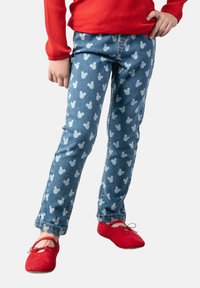 Jasnoniebieskie dżinsy z powtarzającym się białym wzorem głowy Myszki Miki, zestawione z jasnoczerwoną swetrem o prążkowanej strukturze i czerwonymi butami.